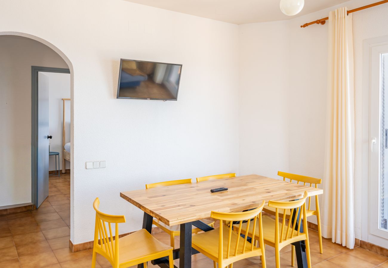 Dining area with wooden table, six yellow chairs and wall TV in a holiday apartment in Llançà, Girona